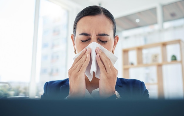 Covid virus and sick with a business woman blowing her nose with a tissue and sneezing while working in her office Allergies sneeze and flu with a female employee suffering from a cold at work