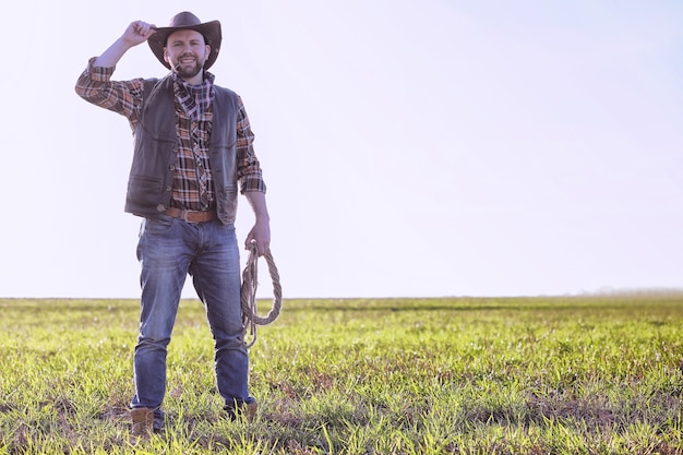 Cowboy in a hat and with a lasso standing in a field at sunset