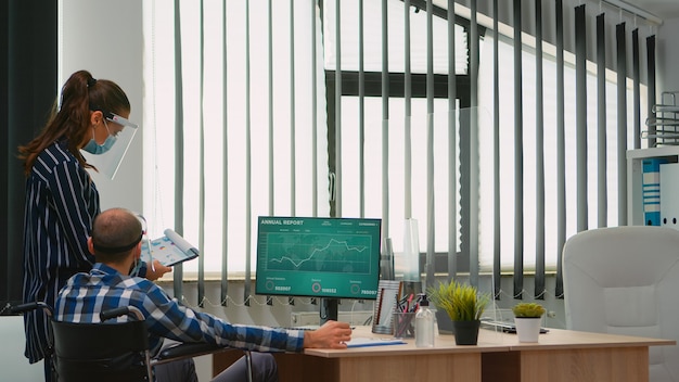 Coworkers with protection face masks working together in workplace during pandemic, businessman moving with wheelchair. Team in new normal business office analysing project graphs typing on computer.