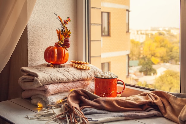 Cozy autumn still life on the windowsill: warm wool sweaters, scarf, pumpkins, maple leaves and a Cup of cocoa with marshmallows and waffles.