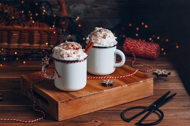 Cozy Christmas composition.Two mug with hot drinks, chocolate with whipped cream and cinnamon stick on a dark wooden . Sweet treats for cold winter days.