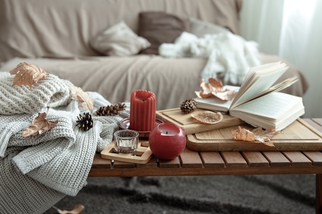 A cozy home composition with candles, a book, knitted sweaters and leaves in the interior of the room.