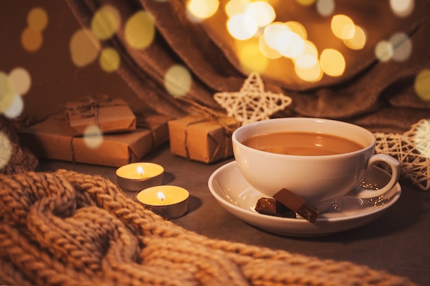 Cozy home still life: white cup with coffee, chunky knit scarf, garland, candles and gifts. The concept of the coming winter, christmas eve and new year. Warm shades, close-up.