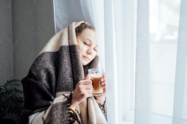 Cozy mood baby its cold outside young woman standing near window with plaid drinking coffee or tea