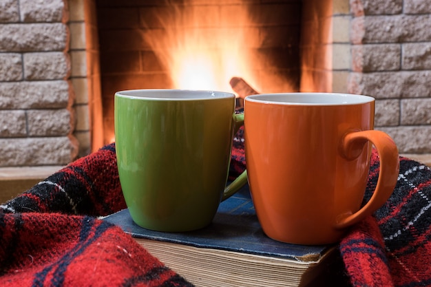 Cozy scene before fireplace with orange and green mugs with tea, and wool scarf.