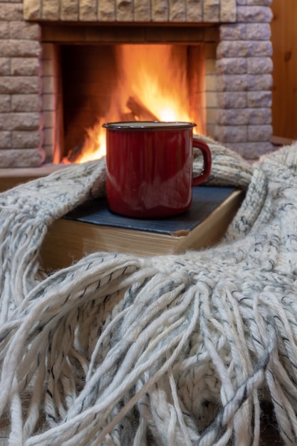 Cozy scene before fireplace with red mug with tea, a book, wool scarf.