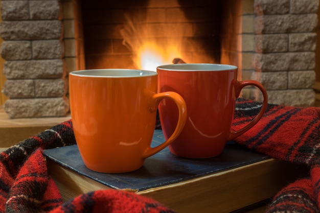 Cozy scene before fireplace with red and orange mugs with tea, a book, wool scarf.