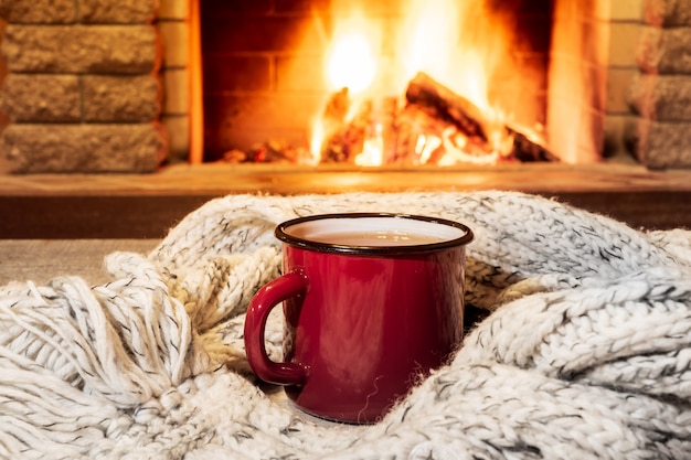 Cozy scene near fireplace with a Red enameled mug with hot tea and cozy warm scarf.