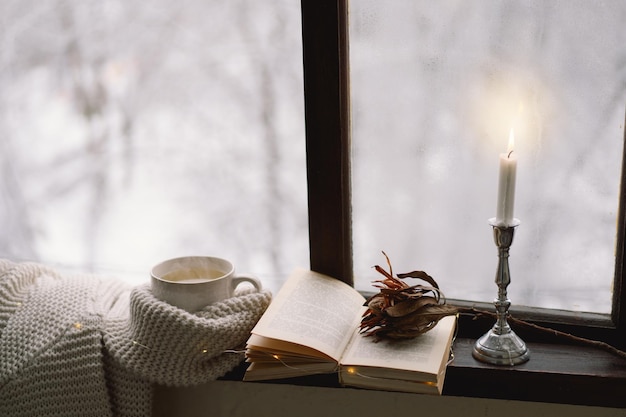 Cozy winter still life Cup of hot tea and an open book with a warm sweater on a vintage wooden windowsill Cozy home concept Sweet home