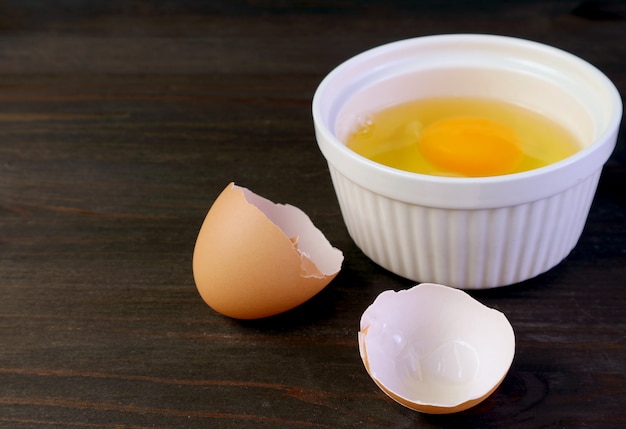 Cracked eggshell on dark brown wooden table with blurry raw egg bowl