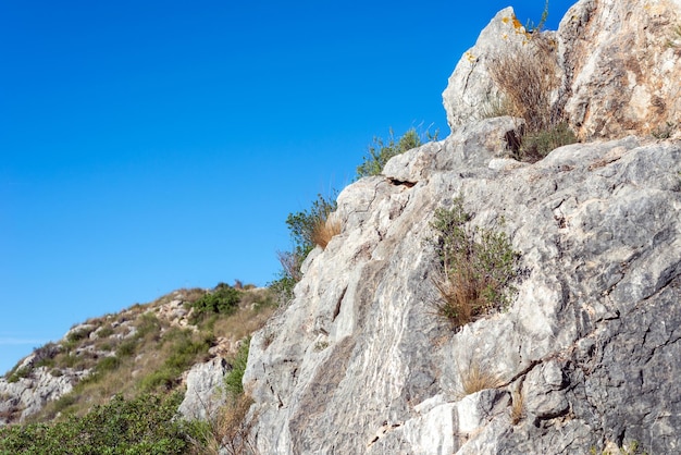 Cracked rock covered with dry grass and plants on a backdrop of a hill and blue sky