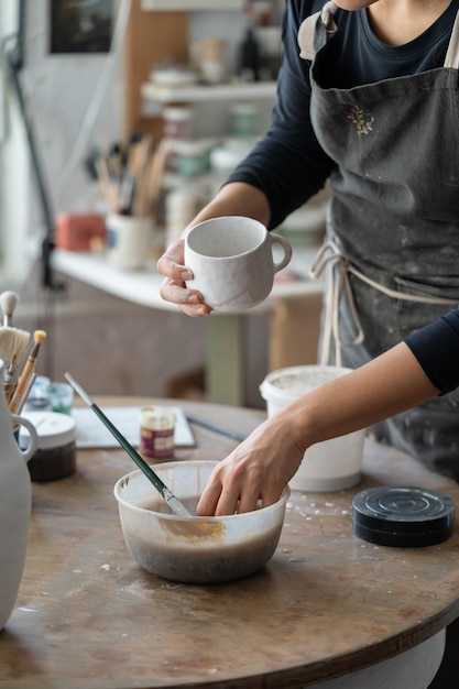 Craftsperson in dirty apron puts hand into bowl with paint to work with handmade pottery in workshop