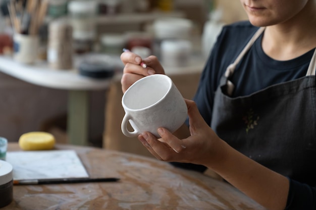 Craftswoman makes sketch of future pattern on white mug to decorate kitchen with clay dish