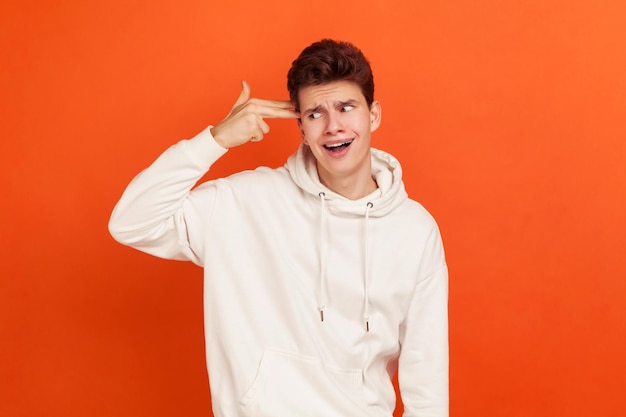 Crazy depressed teenager in casual white hoodie holding fingers near temple imitating gun, suicidal thoughts, paranoia. Indoor studio shot isolated on orange background