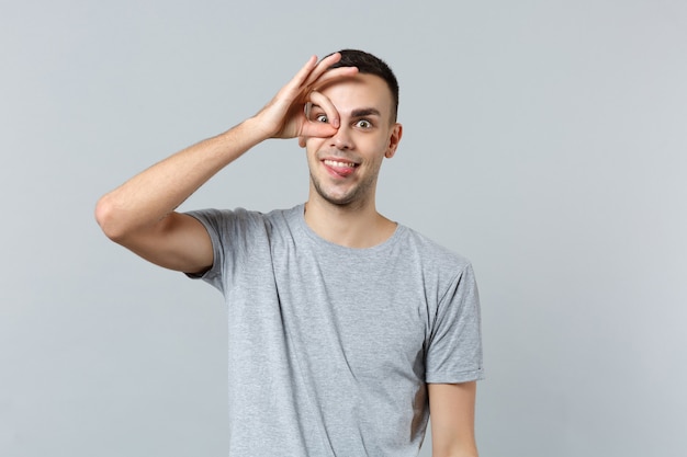 Crazy funny young man in casual clothes showing tongue holding hand near eye imitating glasses or binoculars 