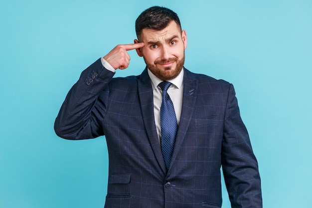 Crazy idea Portrait of man in dark suit showing stupid gesture looking at camera with condemnation and blaming for insane plan dumb suggestion Indoor studio shot isolated on blue background