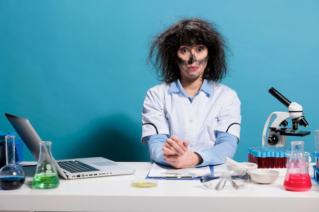 Crazy mad biochemistry expert with messy hair and dirty face sitting at lab desk after experiment explosion. Maniac goofy looking chemist looking dumb while looking at camera on blue background.