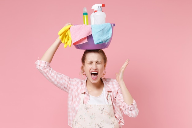 Crazy woman housewife in apron doing housework isolated on pink background. Housekeeping concept. Hold basin with detergent bottles washing cleansers on head keeping eyes closed spreading hand scream.