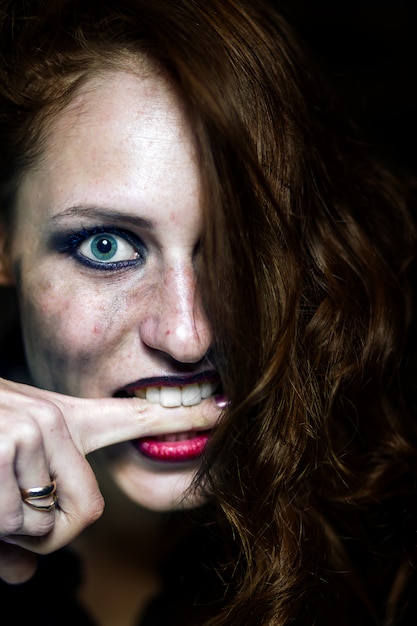 Crazy young girl bites herself on the finger. Emotional portrait. On black background.