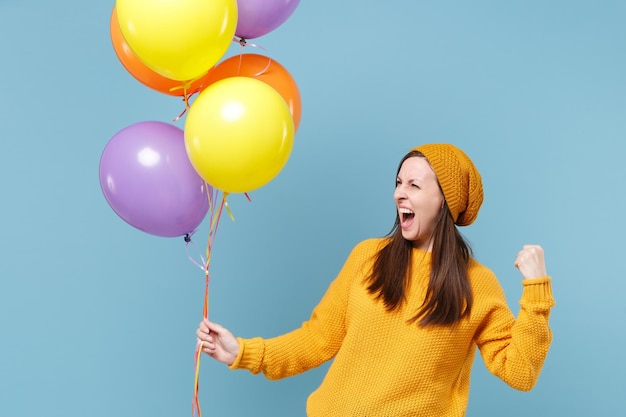 Crazy young woman in sweater hat posing isolated on blue background. Birthday holiday party people emotions concept. Mock up copy space. Celebrating hold colorful air balloons doing winner gesture.