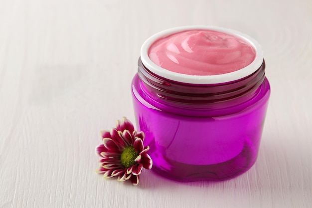 Cream cosmetics for face and body Pink cream and flower in a purple jar on a white wooden table