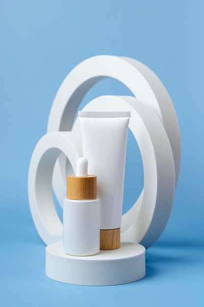 Cream tube mockup and white dropper bottle on  podium  with geometric forms on blue background