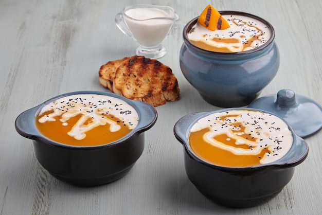 Creamy pumpkin soup in bowls and saucepan with cream black cumin seeds herbs and toasted bread on a blue wooden table