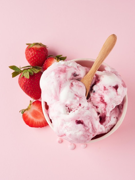 Creamy strawberry ice cream melting in a cup with a wooden spoon on a pink background with fresh strawberries