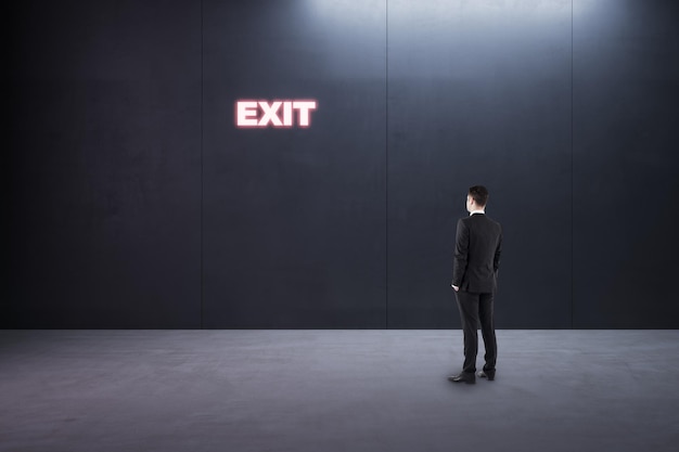 Create your future and best wayout concept businessman back view in empty room looking at glowing neon exit sign on dark wall