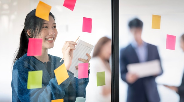 Creative businesswoman reading sticky notes on glass wall with colleague working in background at office