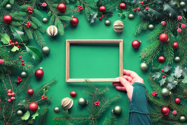 Creative Christmas flat lay on green paper with copy-space. Female hand holding empty frame. Decorative border made of fir and holly twigs, green and red trinkets, dry lime fruit and frosted berries.