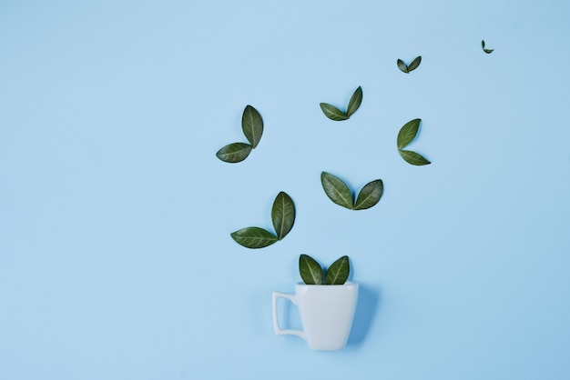 Creative composition. Coffee cup with birds made of natural green leaves on blue background