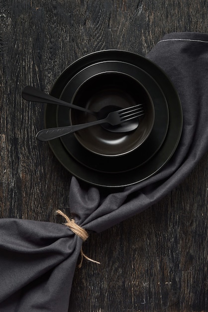 Creative composition from empty black kitchen utensils - dishes and plates with fork and spoon served textile napkin on the same colored background, copy space. Top view.