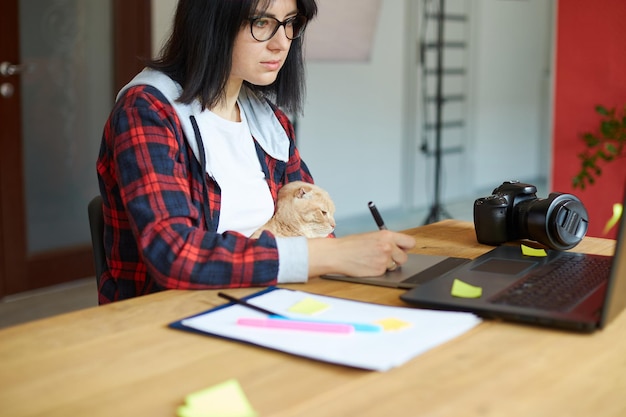 Creative female photographer with cute cat using graphic drawing tablet and stylus pen