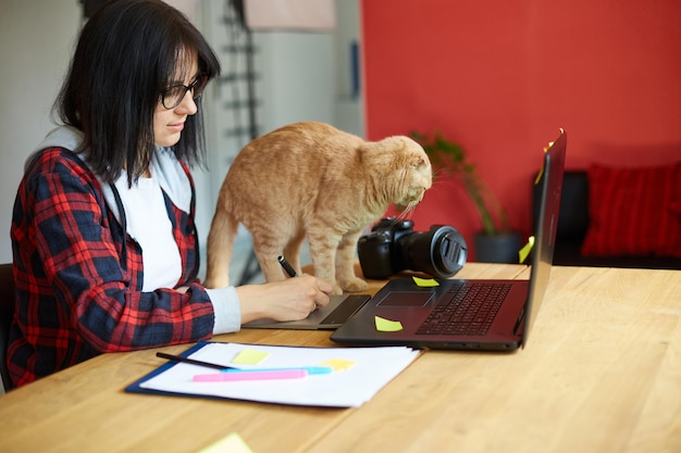 Creative female photographer with cute cat using graphic drawing tablet and stylus pen