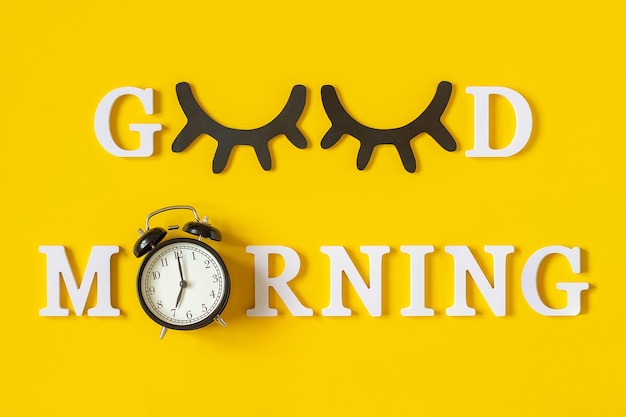 Creative good morning concept White letters black eyeleshes and alarm clock on yellow background Top view Flat lay Greeting card