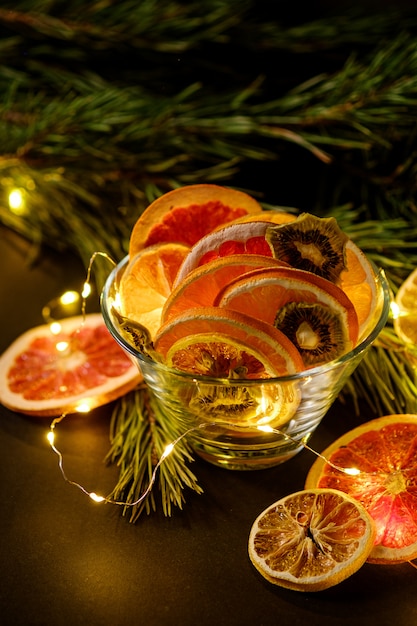 Creative moody holiday Christmas New Year food fruits with dried grapefruit, kiwi, orange and lemon in glass bowl with branch of fir tree with warm led lights, angle view