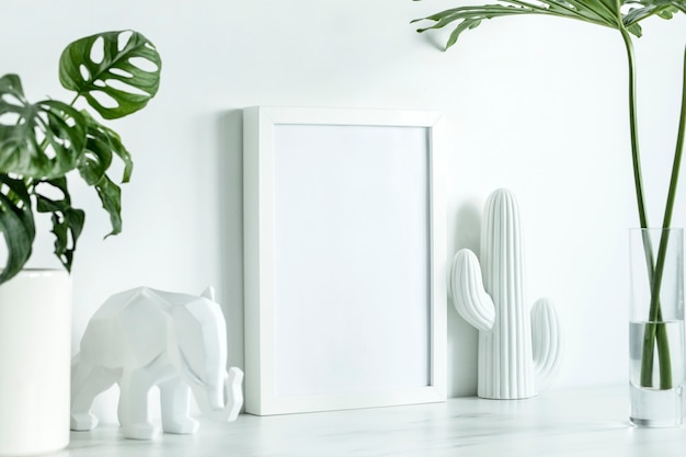 Creative office desk in scandinavian style with white mock up poster frame, white figures of cacti and elephant, leaves in glass vase. White minimalistic concept.