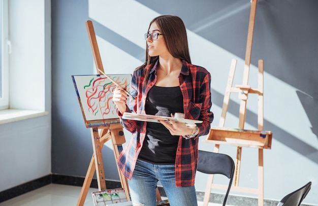 Creative pensive painter girl paints a colorful picture on canvas with oil colors in workshop.