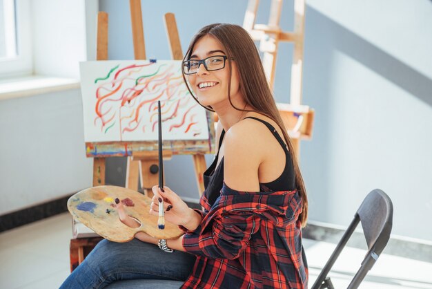 Creative pensive painter girl paints a colorful picture on canvas with oil colors in workshop.