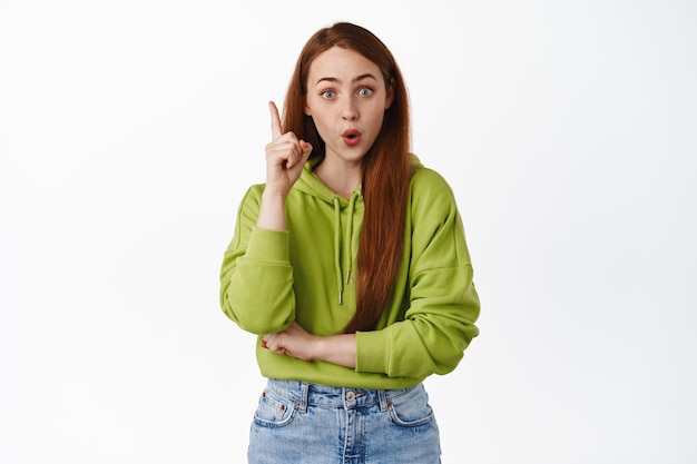 Creative redhead woman have an idea, raising finger in eureka sign and gasp, suggest good plan, have solution, standing against studio background, isolated.