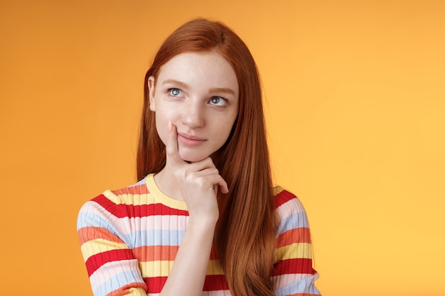 Creative thoughtful young smart redhead female student thinking ideas project look upper left corner dreamy touching lip pondering choice, taking important decision what order, orange background.