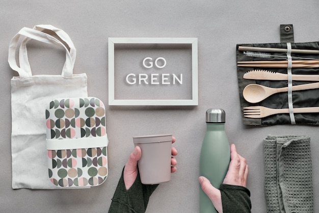 Creative top view, zero waste packed lunch concept. Flat lay, lunch set - bamboo cutlery, lunch box, cotton bag and hand with coffee-to-go cup on craft paper. Frame with text "Go green".