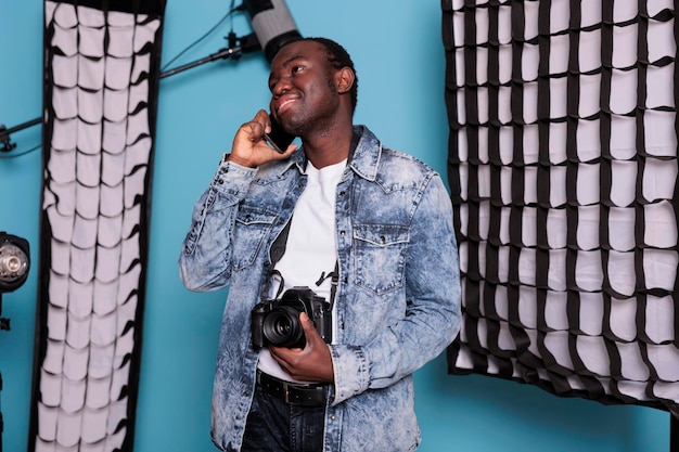 Creative video producer on blue background with spotlight while having a phone call. Professional photographer talking on smartphone while standing in studio having softboxes and production equipment.