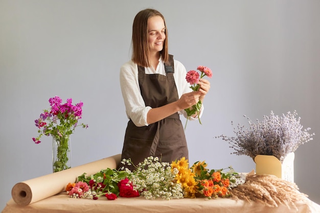 Creative workspace decor Market for organic blooms Attractive happy woman florist creating beautiful bouquet at workplace in flower shop standing isolated over gray background