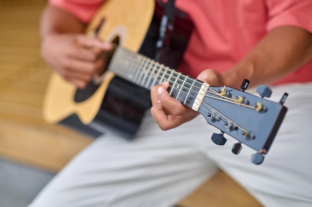 Creativity, music. Hands of dark skinned man in red tshirt and white trousers playing guitar outdoors on fine day, without face