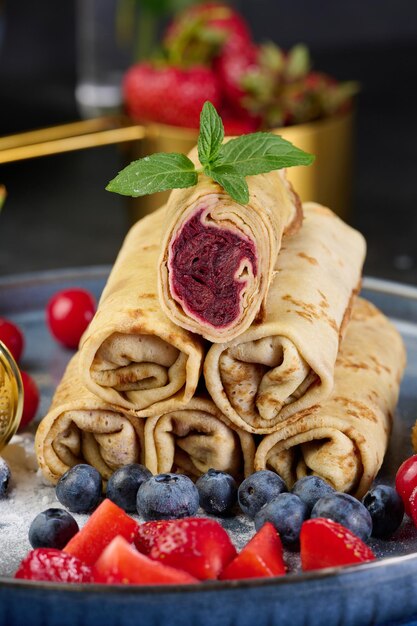 Photo crepes stacked in a pile one of which is cut and filled with berry filling the plate is also decorated with blueberries strawberries and cherries