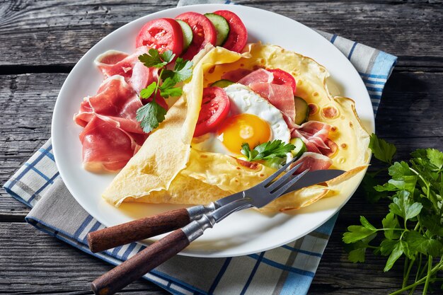 Crepes with fried egg, Jamon slices, fresh tomatoes and cucumber fillings on a white plate on an old wooden table, view from above