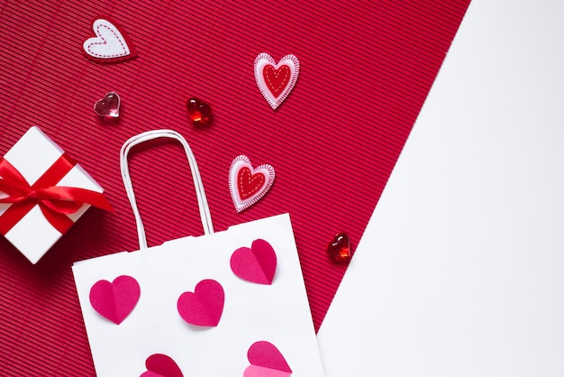 On a crimson and white background, a gift box with a pink bow, a white paper bag, crimson glass hearts hearts