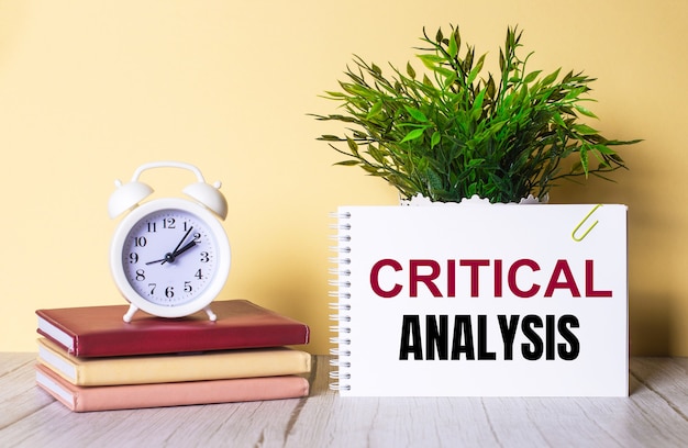 CRITICAL ANALYSIS is written in a notebook next to a green plant and a white alarm clock, which stands on colorful diaries